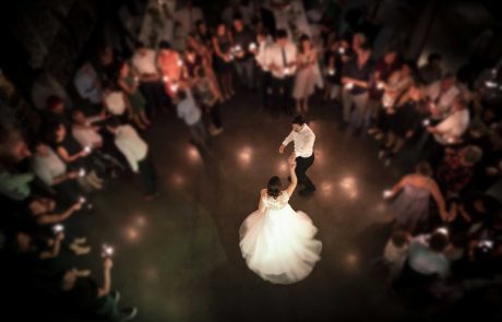 A bride and groom dance at the center of a circle of onlookers in a dimly lit venue, with guests holding small lights.