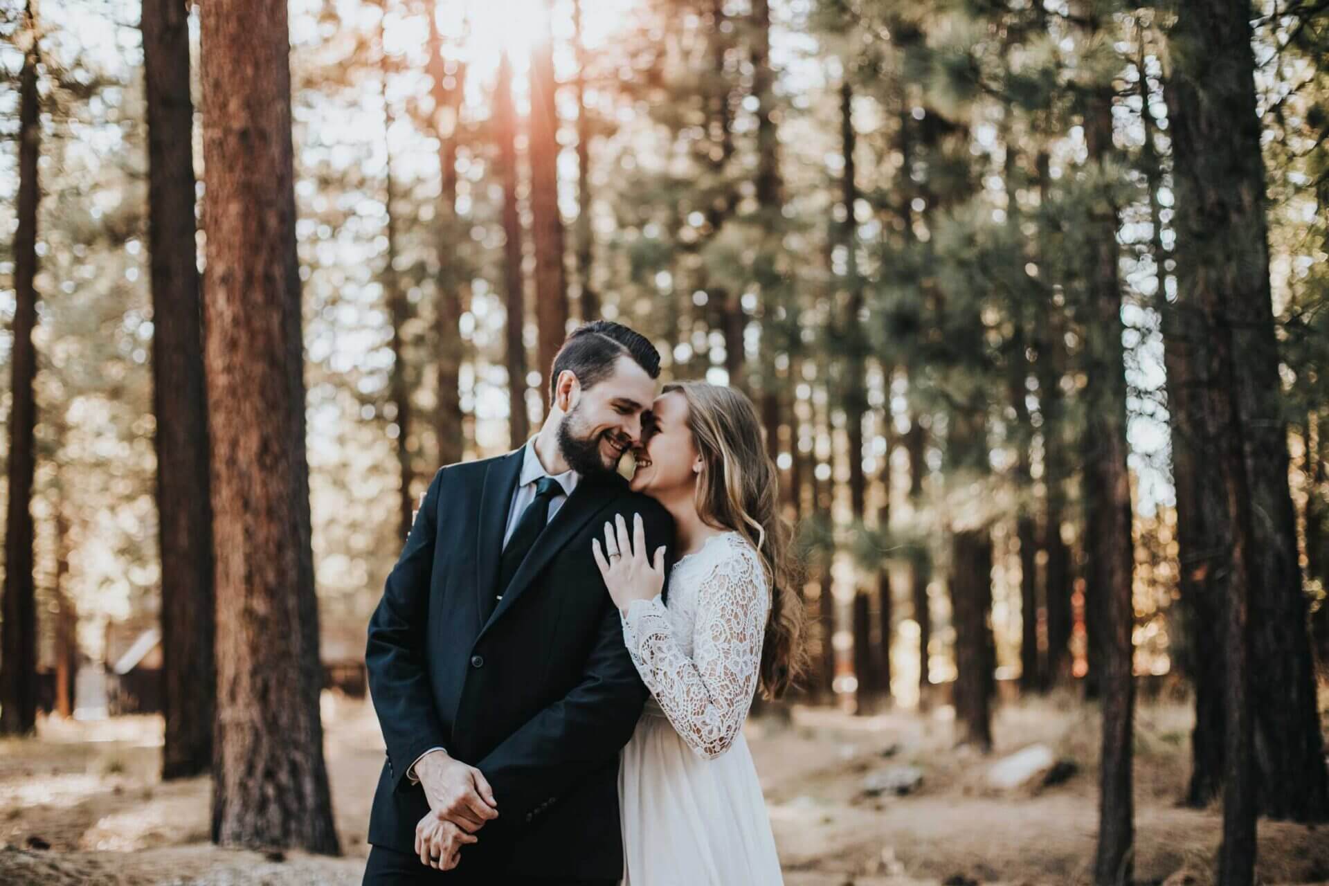 A couple dressed formally stand close together in a forest, smiling and embracing each other with sunlight shining through the trees.