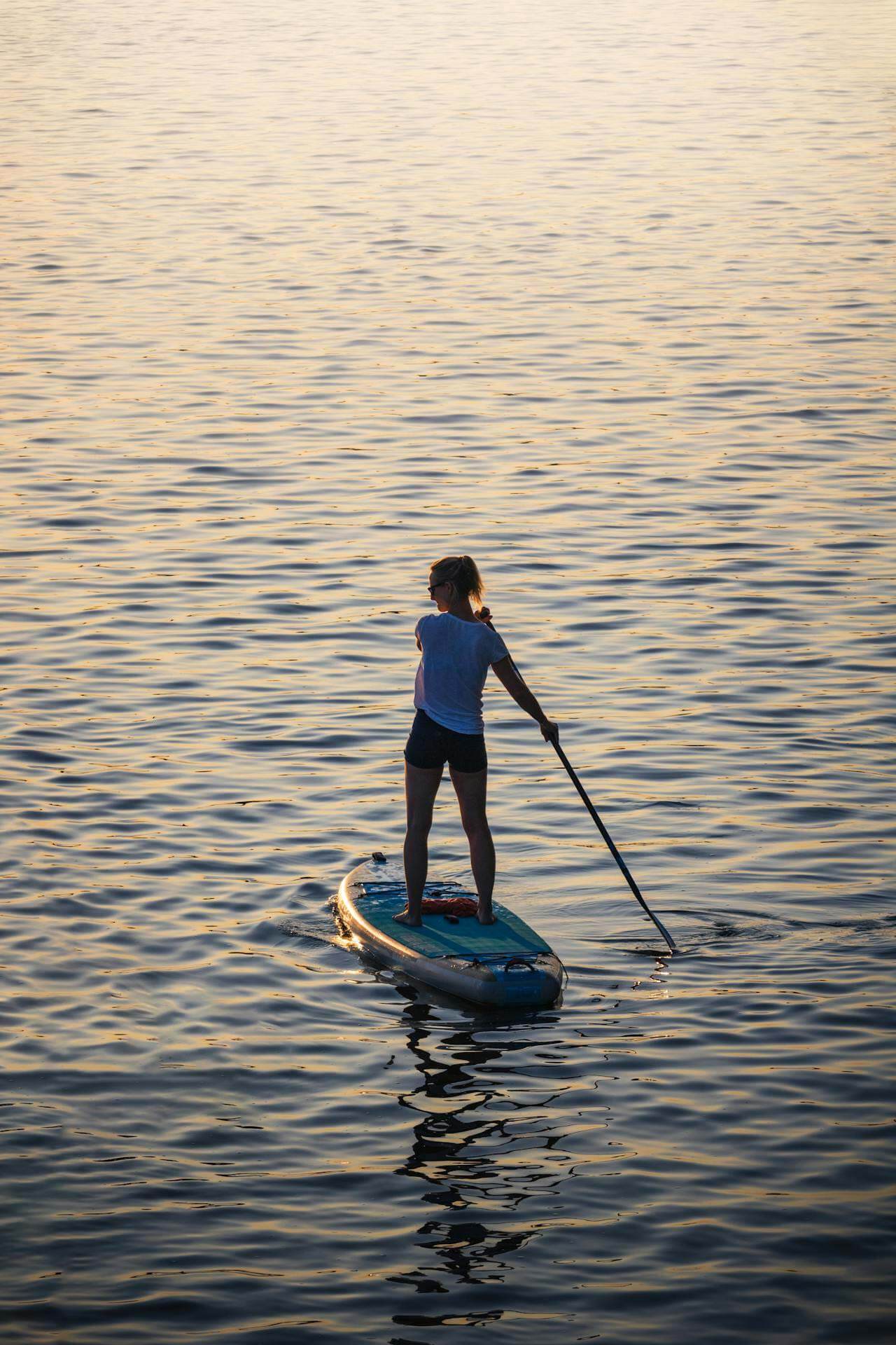 A person stands on a paddleboard and uses a paddle to move across calm water during sunset or sunrise.