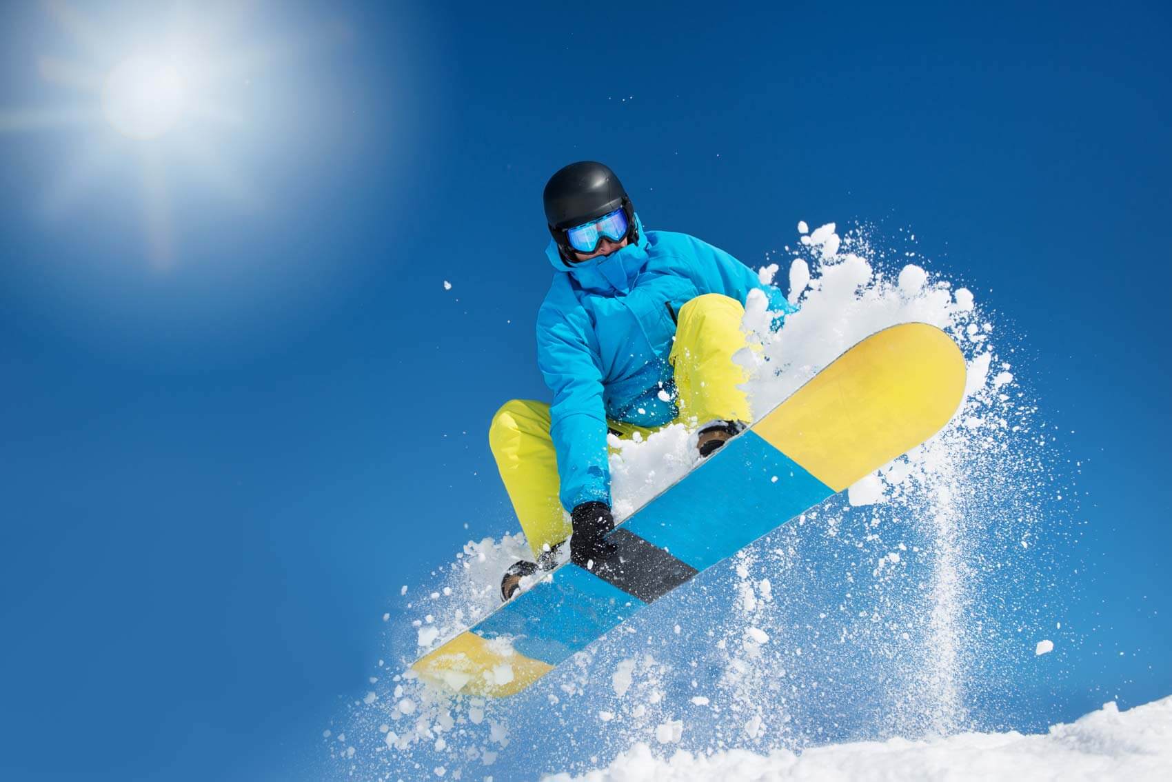Snowboarder in blue and yellow gear jumps in the air, kicking up snow, with a clear blue sky and bright sun in the background.