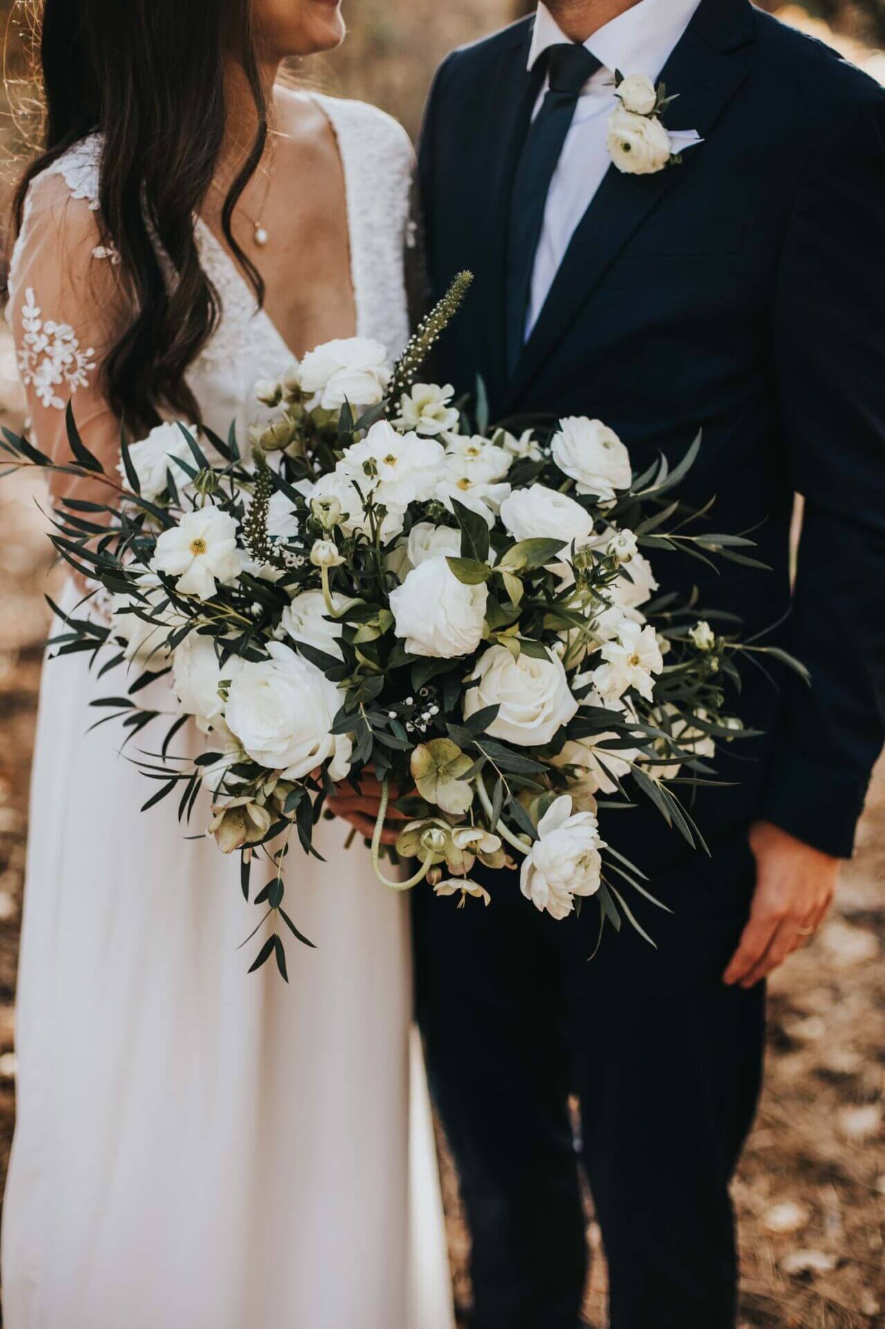 A bride in a white dress holding a large bouquet of white flowers stands next to a groom in a dark suit with a white boutonniere. Their faces are partially out of frame.