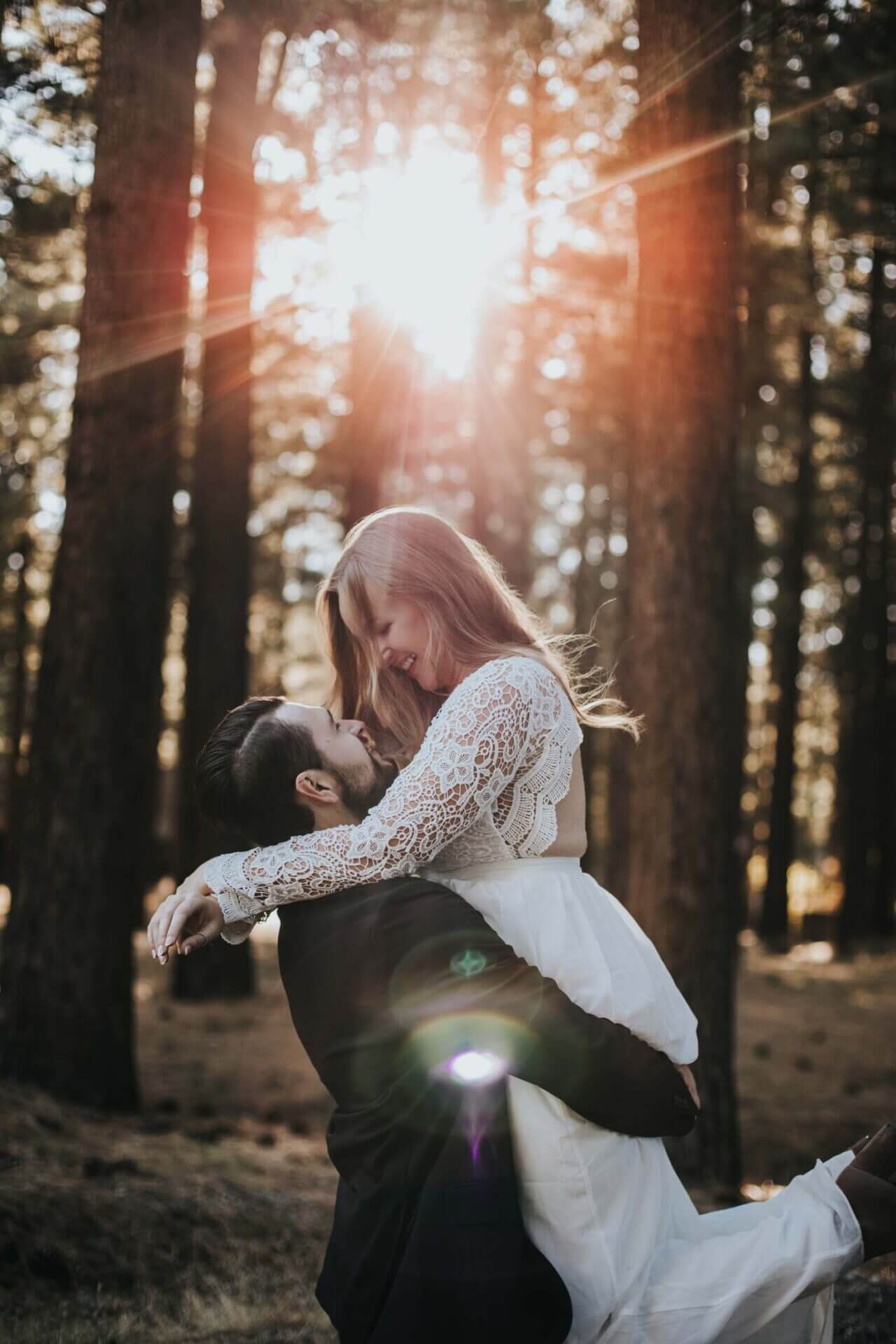 A man lifts a woman in a white lace dress in a forest, with sunlight streaming through the trees behind them.