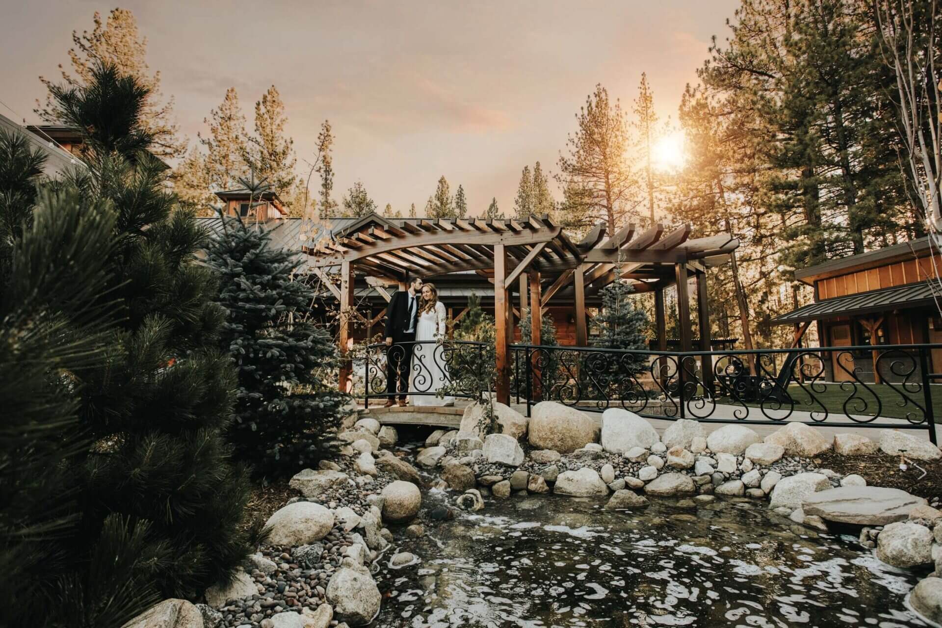 A couple stands together on a wooden bridge under a pergola, surrounded by pine trees and rocks, with the sun setting in the background.