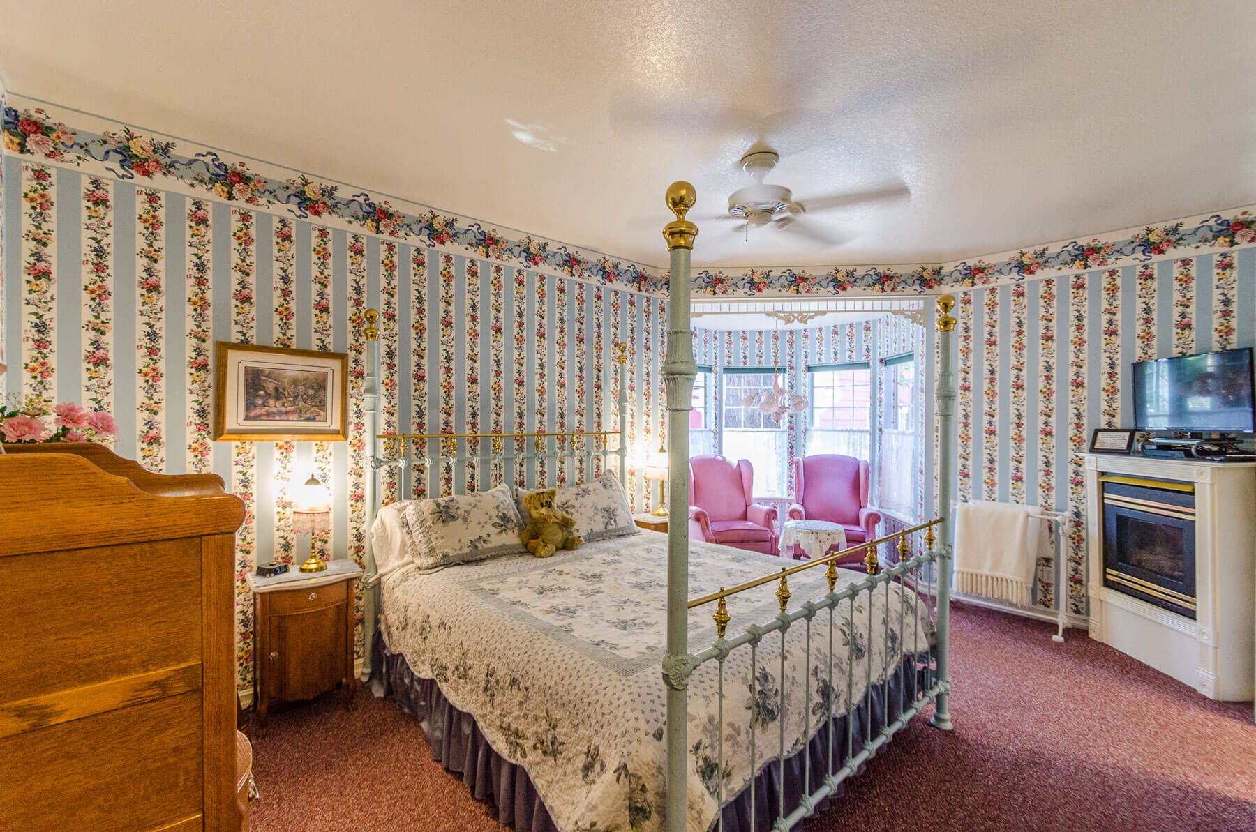 Victorian-style bedroom with floral wallpaper, an iron bed, two pink armchairs by a window, a fireplace, TV, and a wooden dresser.