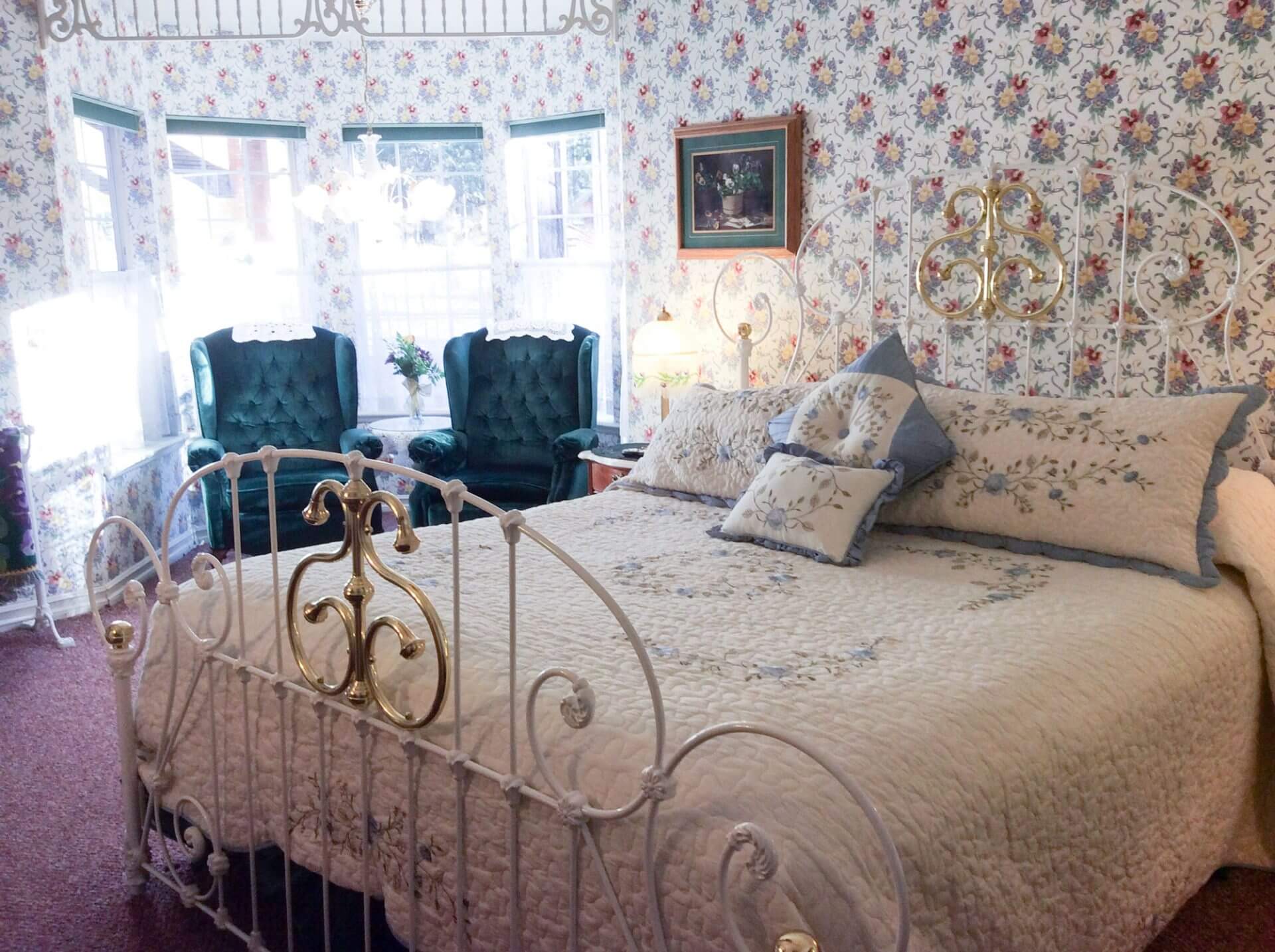 A bedroom with a white ornate metal bed, floral bedding, floral wallpaper, two green armchairs by bay windows, and framed art on the wall.
