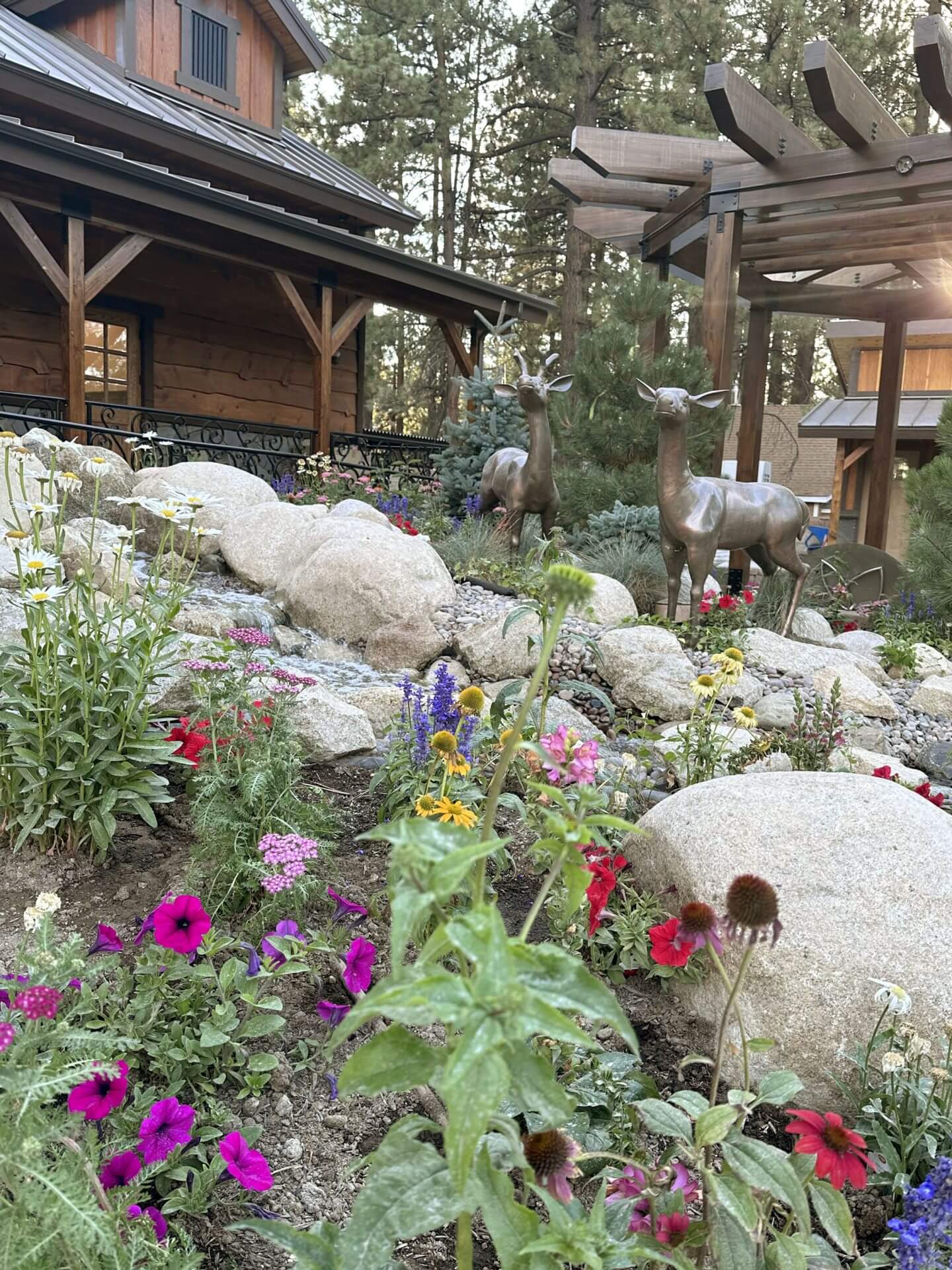 A landscaped garden with colorful flowers, large rocks, and two deer statues in front of a wooden house surrounded by trees.
